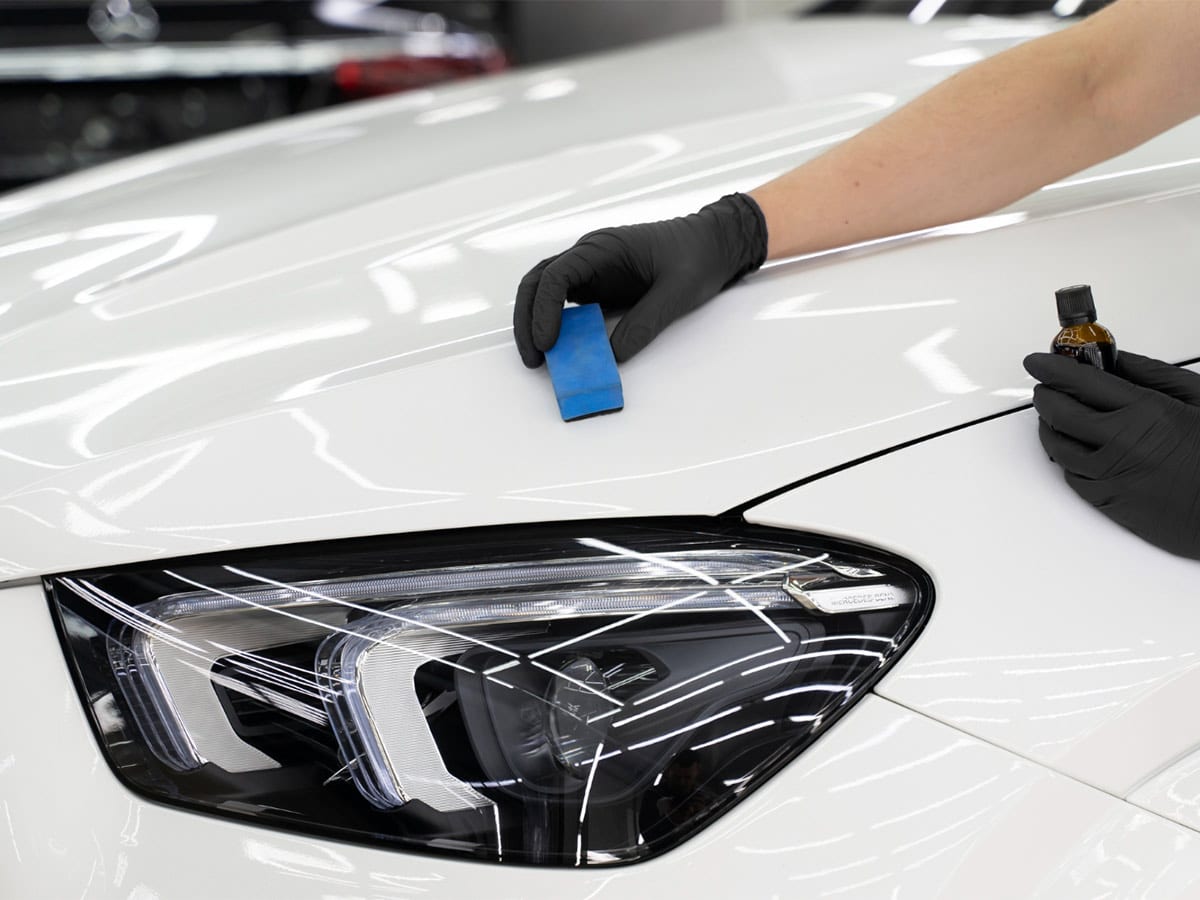 A person in black gloves cleans a white car, applying a ceramic/glass coating to protect the paint.