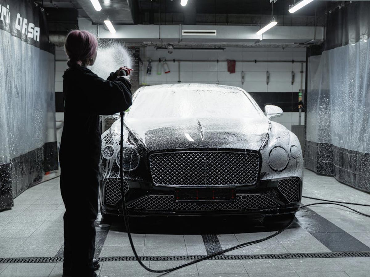 A woman is engaged in car detailing, washing a Bentley in a well-lit garage setting.