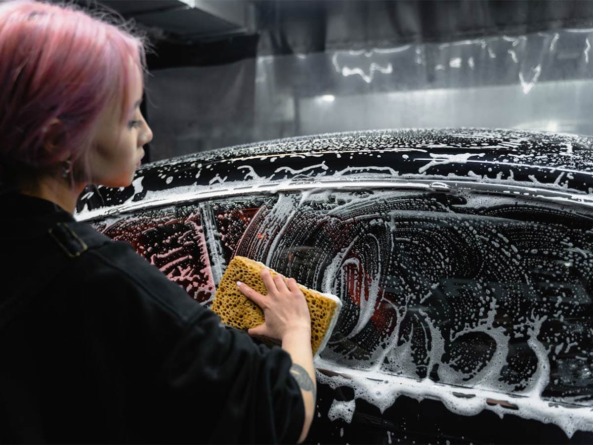 A auto cleaner woman with pink hair cleaning car windows with the glass cleaner using a sponge.