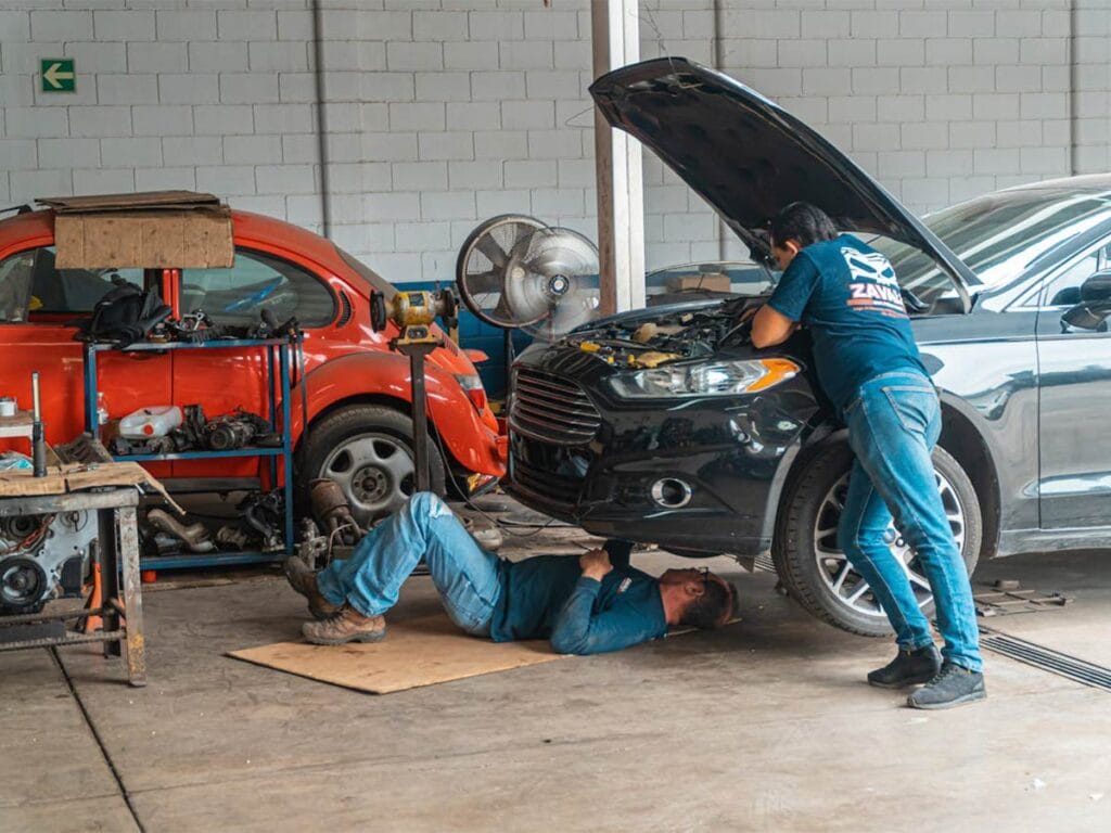 Two men, dressed in work clothes, repair a car inside a well-lit garage, surrounded by tools and equipment.